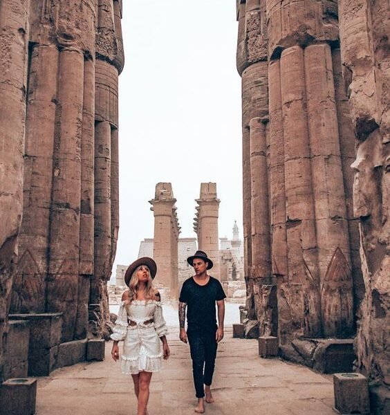 A couple walking barefoot among ancient stone columns at a historical site.