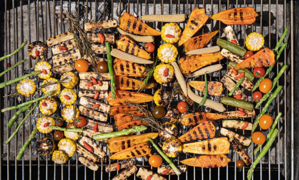 Grilled vegetables including corn, carrots, asparagus, and tomatoes on a BBQ grill.