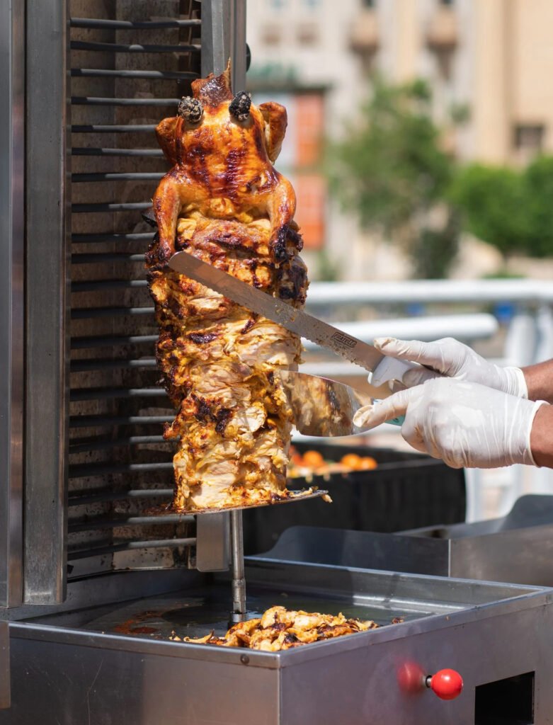 Rotisserie chicken and shawarma being sliced by a person in gloves on a street stand.