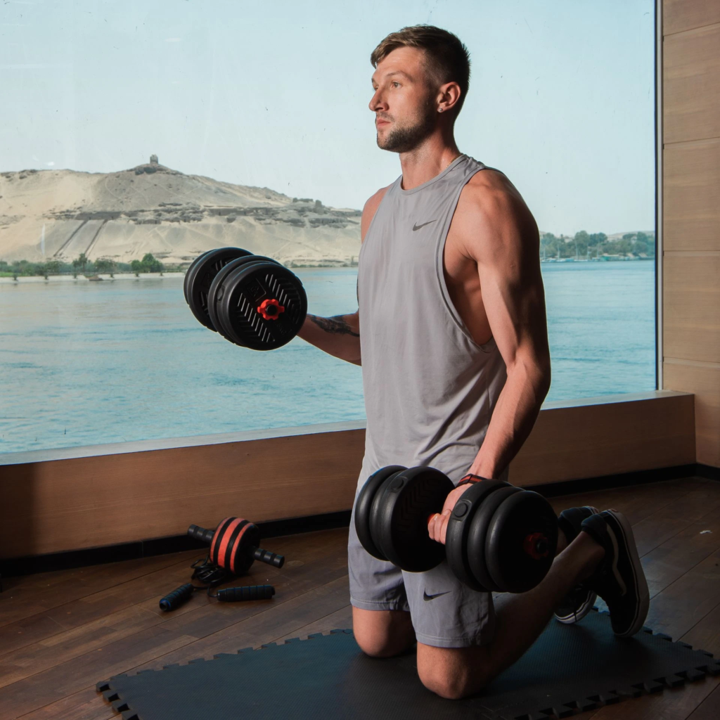 Man in gym clothes doing a kneeling dumbbell lift with a scenic river background.