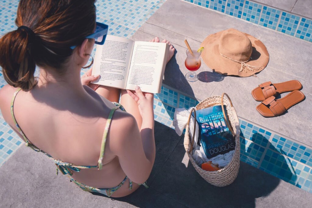 Person reading by the poolside with a hat, cocktail, sandals, and bag nearby.