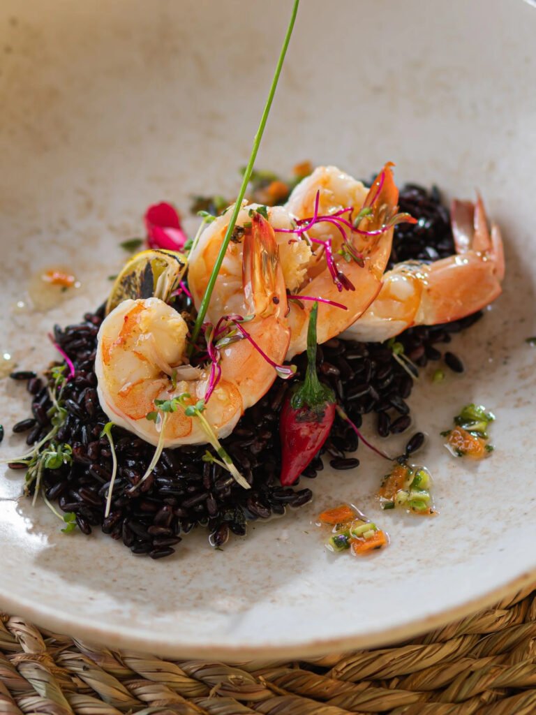 Gourmet dish with prawns on black rice and colorful garnishes in a ceramic bowl.