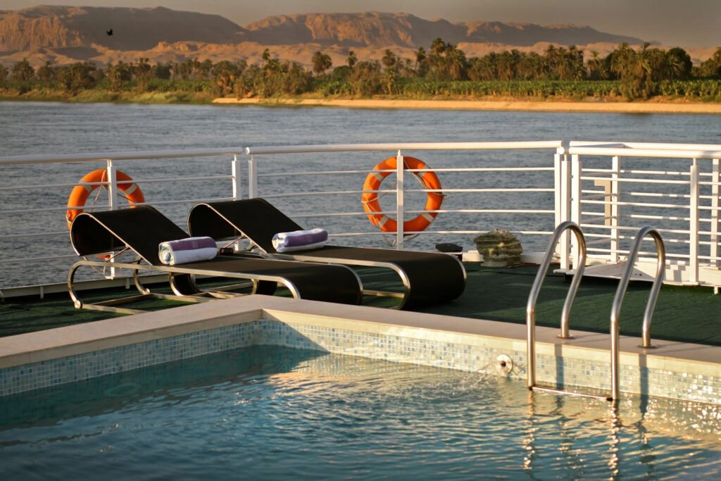 Cruise ship deck with sun loungers by a small pool, overlooking a river and distant mountains.