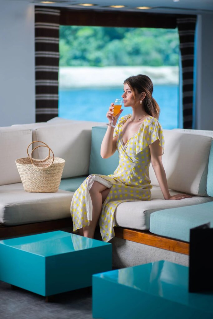 Woman in yellow dress sitting on a sofa, holding a drink, with a straw bag beside her.
