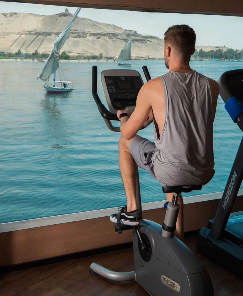 Man exercising on a stationary bike by a window overlooking a river with sailboats.