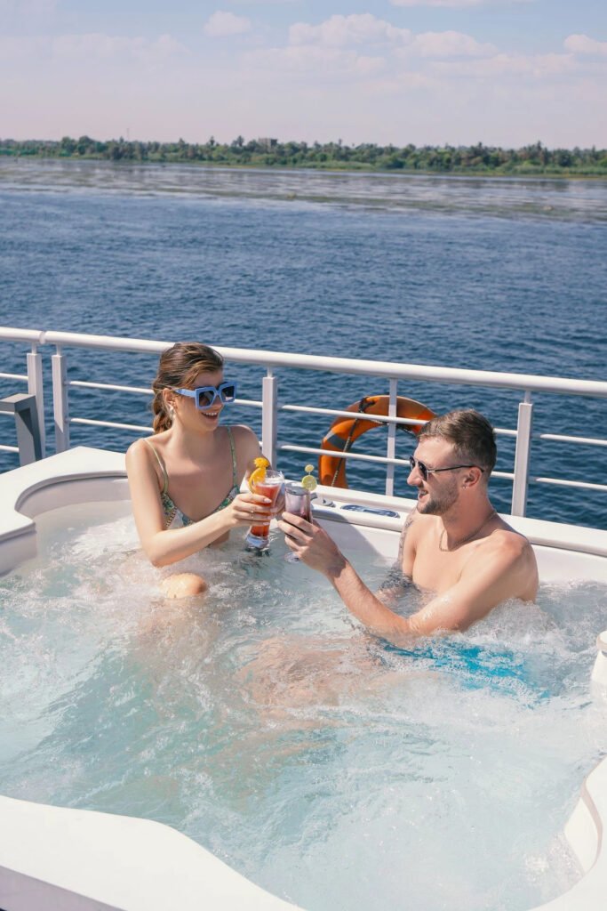 Two people toasting drinks in a hot tub on a boat with a river in the background.