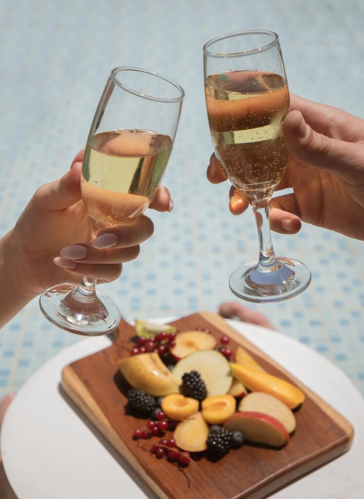 Two hands toasting with champagne over a fruit platter by a pool.