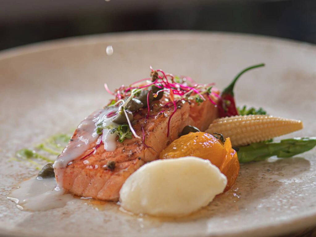 Grilled salmon garnished with microgreens, sauce, and sides of baby corn and citrus on a plate.