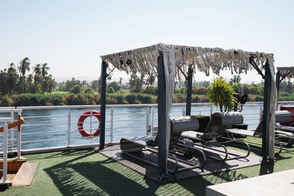 Deck of a boat with sun loungers and a thatched canopy, overlooking a river and palm trees.