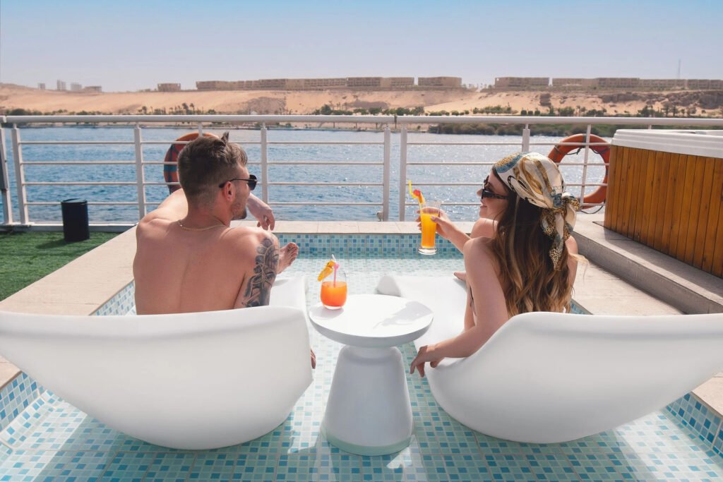 Two people relaxing in deck chairs by a pool, overlooking a river and sandy hills.