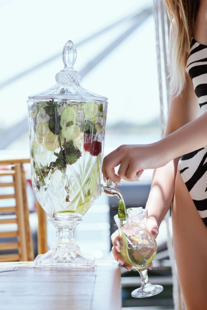 Person dispensing infused water from a large glass dispenser into a cup.