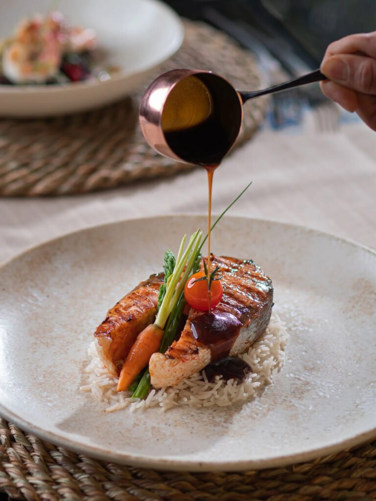 hand pouring sauce over grilled salmon on rice with vegetables.